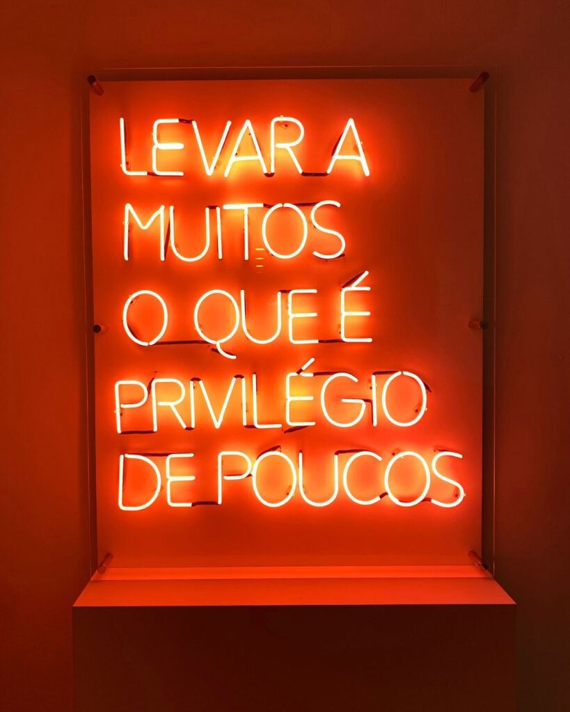 Frase em neon vermelho: “Levar a muitos o que é privilégio de poucos”. A mensagem traduz a missão da empresa e seu foco no desenvolvimento de profissionais por meio da democratização de oportunidades.
