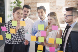 Grupo de profissionais colaborando entre si durante uma trilha de desenvolvimento de profissionais, utilizando post-its coloridos em um quadro, promovendo a troca de ideias e o aprendizado coletivo.