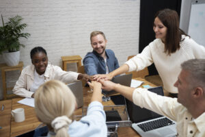Equipe diversa reunida em um ambiente de escritório, unindo as mãos no centro da mesa em um gesto de colaboração. A imagem representa a superação da cultura de silos nas empresas, com profissionais engajados trabalhando juntos de forma integrada.