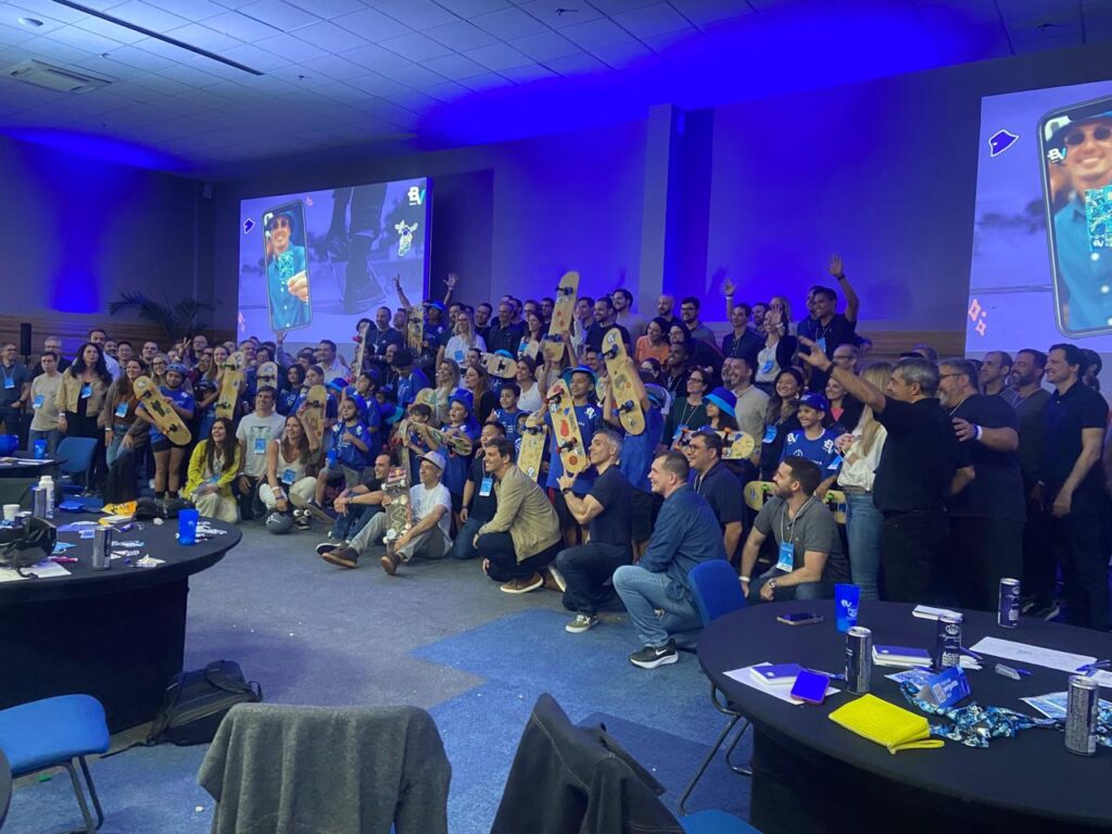 Imagem de um evento de Team Building de fim de ano, com um grande grupo de colaboradores reunidos em uma sala de conferências. A equipe segura pranchas de skate personalizadas, celebrando em um ambiente descontraído com um toque de estratégia e integração. O espaço é iluminado em tons de azul, com projeções de telas ao fundo e clima de entusiasmo e união para o próximo ciclo.