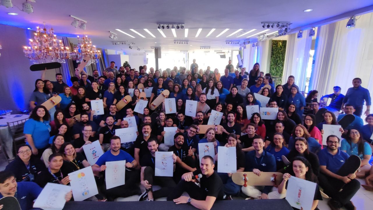 Imagem de um evento de Team Building de fim de ano, com um grande grupo de funcionários sorrindo enquanto seguram pranchas de skate personalizadas e desenhos. O ambiente é descontraído e festivo, com decoração iluminada e um clima de celebração de propósito. A equipe está unida, refletindo o fortalecimento e engajamento da empresa para o novo ciclo.