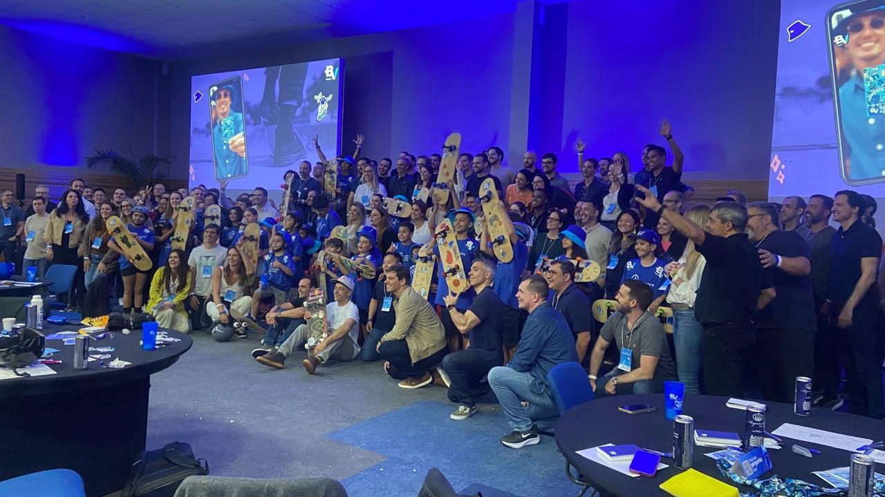 Imagem de um evento de Team Building de fim de ano, com um grande grupo de colaboradores reunidos em uma sala de conferências. A equipe segura pranchas de skate personalizadas, celebrando em um ambiente descontraído com um toque de estratégia e integração. O espaço é iluminado em tons de azul, com projeções de telas ao fundo e clima de entusiasmo e união para o próximo ciclo.